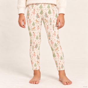 May include: Cream-colored leggings featuring a repeating pattern of nutcrackers, ballerinas, rocking horses, Christmas trees, and candy canes in soft shades of pink, green, and brown. The leggings have a high waistband and a fitted silhouette.