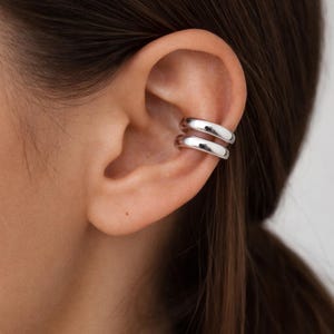 Peut inclure: Une boucle d'oreille de type ear cuff argentée présentant un design à double anneau avec deux cercles métalliques lisses, polis et arrondis qui entourent le cartilage de l'oreille. Le bijou possède une finition métallique réfléchissante.