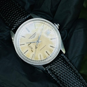 May include: A vintage Longines automatic wristwatch with a round silver-tone case and a textured black strap. The watch face is cream-colored with visible patina, featuring silver hour markers, a date window at the three o'clock position, and the text Longines and Automatic printed on the dial.