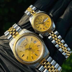 May include: Two West End Watch Co. Sowar wristwatches with gold-toned dials and fluted bezels. Both feature two-tone silver and gold-colored metal link bracelets. The larger watch displays the day and date, while the smaller watch shows the day and date.
