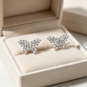 Puede incluir: Un par de pendientes de botón con forma de mariposa en tono plateado, cada uno con múltiples piedras transparentes de talla marquesa que forman las alas. Los pendientes se muestran lado a lado, destacando un acabado metálico y piedras blancas brillantes.