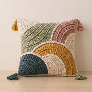 May include: A square crochet throw cushion featuring a textured geometric pattern of concentric semicircles in cream, sage green, dusty rose, mustard yellow, and dark teal. The cushion is finished with four matching tassels at each corner.