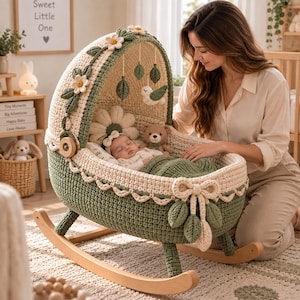 Peut inclure: Un berceau pour bébé en crochet vert sauge et crème avec un bord festonné, un nœud décoratif et un auvent orné de fleurs en tissu et de décorations suspendues en forme de feuilles et d'oiseaux sur des pieds à bascule en bois.