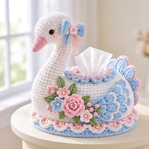 May include: A white crocheted swan tissue box cover featuring a pink beak, a blue and pink bow on its head, and a decorative arrangement of pink and blue crocheted flowers with green leaves on its side. The swan has blue and pink tail feathers.