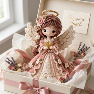 May include: A crocheted angel doll with brown hair, a gold halo, and white wings, wearing a pink and white dress adorned with small flowers. The doll holds a gold heart and is presented in a gift box with a card reading For a special Angel.