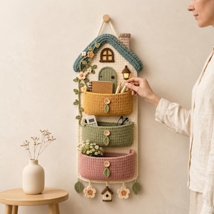 May include: A wall-hanging crochet organizer shaped like a house with three stacked pockets in mustard yellow, sage green, and dusty pink. The design features a blue roof, a small arched door, a lantern, and decorative floral and leaf crochet accents.