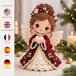 May include: A crocheted angel doll with brown hair styled in an updo, adorned with a floral crown. The doll wears a deep red dress with a white skirt, green leaf accents, and small red flowers. It has white wings and holds a small white heart.