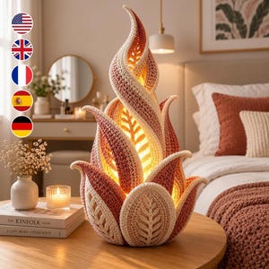 Può includere: Una lampada da tavolo decorativa a forma di fiamma, realizzata in filato lavorato all'uncinetto nei toni del crema, rosa tenue e rosa antico. La lampada emette una luce calda e interna attraverso i punti testurizzati a foglia.