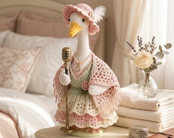 May include: A crocheted goose figurine wearing a pink hat with a white feather, a pearl necklace with a pendant, and a layered dress in shades of pink, cream, and sage green. The goose holds a gold-colored vintage-style microphone stand.