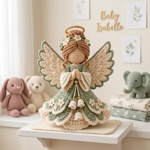 May include: A crocheted angel figurine with light brown yarn hair, a gold halo, and large textured wings. The angel wears a sage green and cream dress adorned with small white flowers and dangling bell-shaped accents, with hands clasped in prayer.