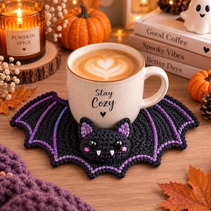 Puede incluir: Una taza de cerámica moteada con el texto Stay Cozy y un icono de corazón, llena de arte latte, sobre un posavasos de ganchillo negro con forma de murciélago con detalles de alas moradas, mejillas rosadas y colmillos blancos.