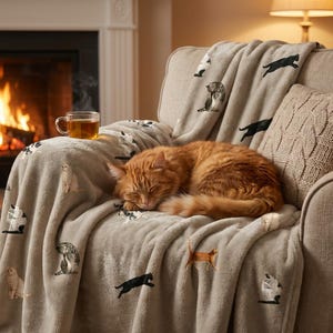 May include: A soft, light gray throw blanket featuring a repeating pattern of various cat illustrations in different poses. A ginger cat is curled up asleep on the blanket, which is draped over a beige armchair next to a glass mug of tea.