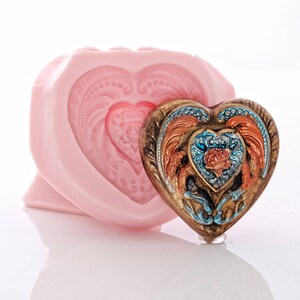 May include: A pink silicone heart-shaped mould with a detailed design and a matching heart-shaped resin piece with a gold, blue, and orange design.