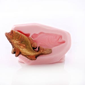 May include: A pink silicone mold with a gold-colored fish figurine. The mold is shaped like a fish and has a detailed impression of a fish on the inside.