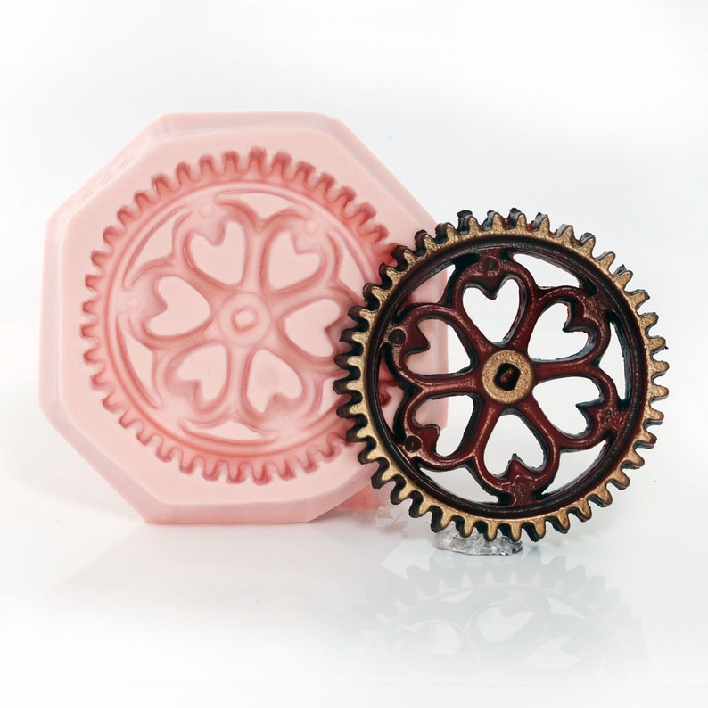 May include: A pink silicone mold with a detailed heart-shaped design and a brown metal gear with a similar heart-shaped design.