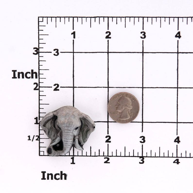 Puede incluir: Una figurita de cabeza de elefante gris con una superficie texturizada. La figurita mide aproximadamente 2,5 cm de alto y se muestra junto a una moneda de 25 centavos de d&oacute;lar estadounidense para comparar el tama&ntilde;o.