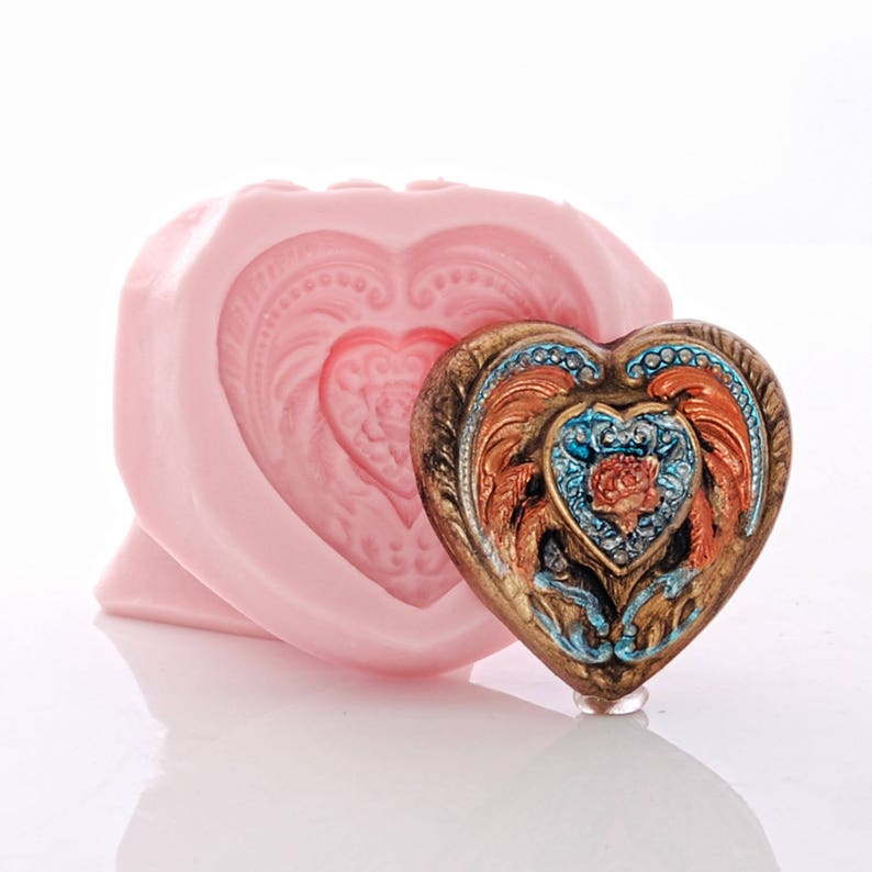 May include: A pink silicone heart-shaped mould with a detailed design and a matching heart-shaped resin piece with a gold, blue, and orange design.