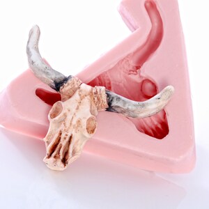 May include: A white ceramic bull skull with horns, resting on a pink silicone mold. The mold is shaped like a bull's head, with a hollow space for the skull to fit into.