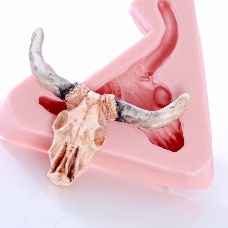 May include: A white ceramic bull skull with horns, resting on a pink silicone mold. The mold is shaped like a bull's head, with a hollow space for the skull to fit into.