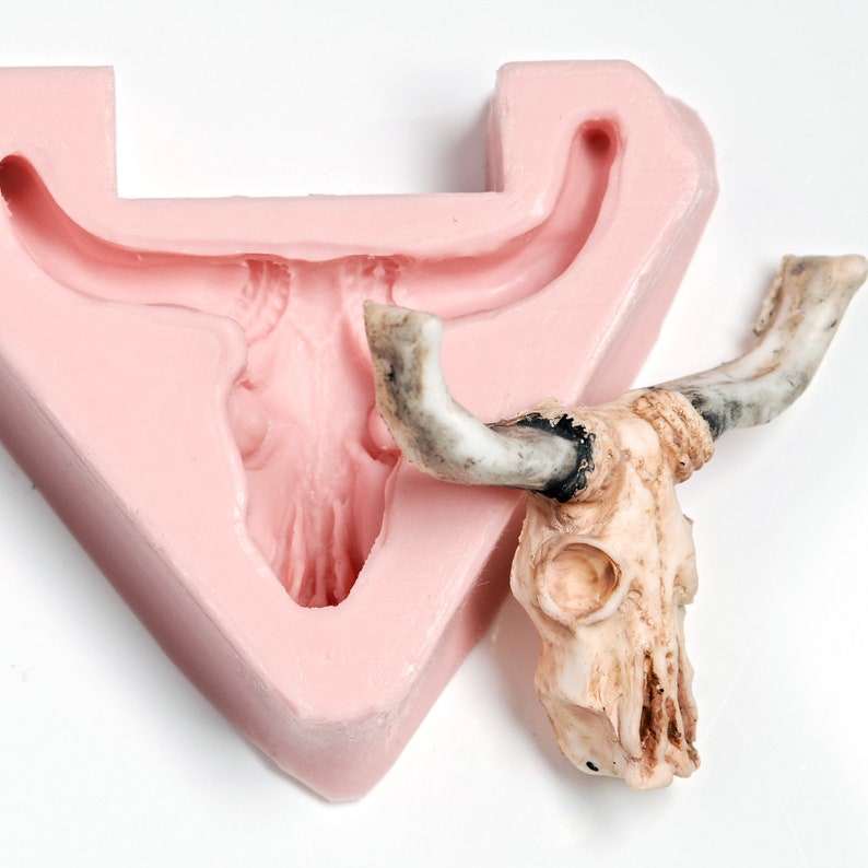 May include: A pink silicone mold of a bull skull with a white and black bull skull sitting next to it.
