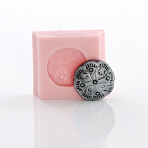 Op de afbeelding: Een roze siliconen mal voor het maken van een decoratieve koek of taarttopper. De mal is gevormd als een ronde koek met een gedetailleerd bloemmotief. Een zilverkleurige koek wordt naast de mal getoond.