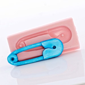 Peut inclure: Une épingle à nourrice en plastique bleue avec un moule en silicone rose. Le moule est en forme d'épingle à nourrice.