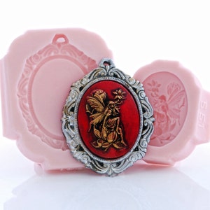 May include: A silver-toned oval frame with an ornate design surrounds a red cameo featuring a gold fairy with wings outstretched. The cameo is set against a pink background.