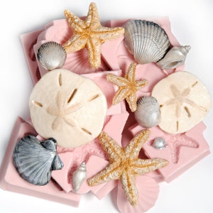 Könnte beinhalten: Eine Sammlung von rosa Silikonformen in Form von Muscheln, Seesternen und Sanddollars. Die Formen sind mit weißer, silberner und goldener Harz gefüllt.