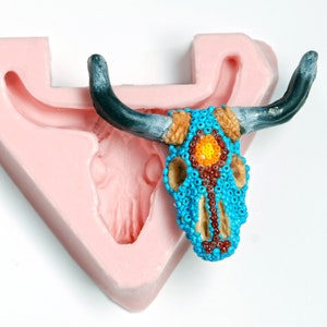 May include: A pink silicone mold shaped like a bull skull with a beaded bull skull inside. The beaded skull is blue with orange and red accents.
