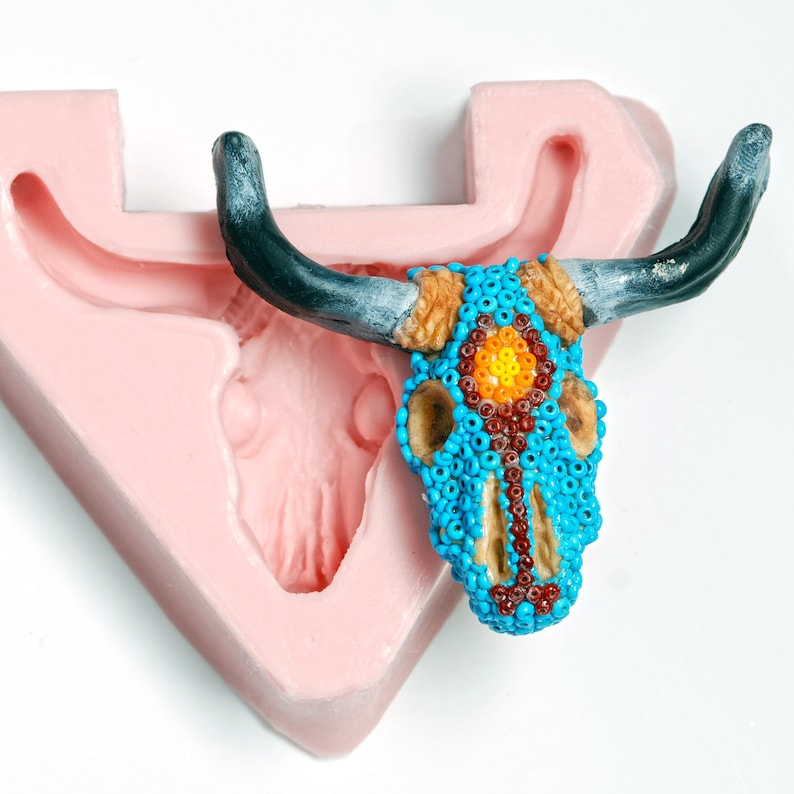 May include: A pink silicone mold shaped like a bull skull with a beaded bull skull inside. The beaded skull is blue with orange and red accents.