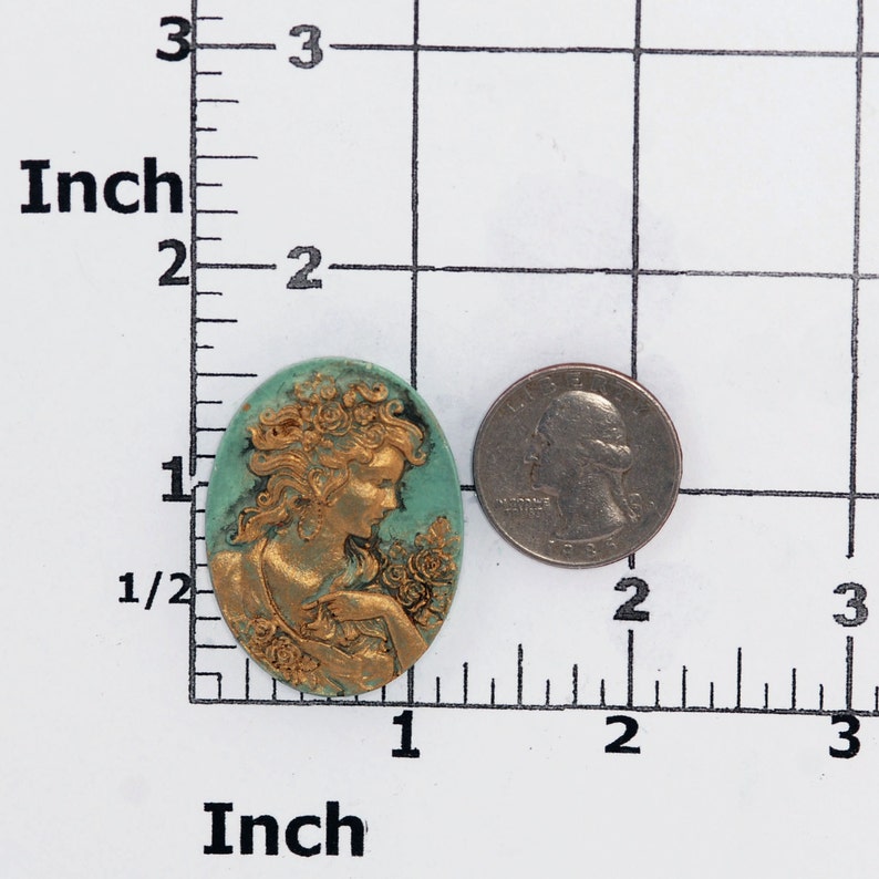 May include: An oval cameo featuring a gold-toned relief of a person with flowing hair and floral accents against a teal background, positioned next to a US quarter on a grid marked in inches for scale reference.