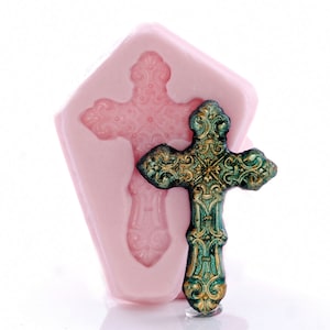 May include: A pink silicone mould for making a decorative cross. The mould is shaped like a coffin and has a detailed design. A finished cross is shown next to the mould. The cross is made of metal and has a green and gold finish.
