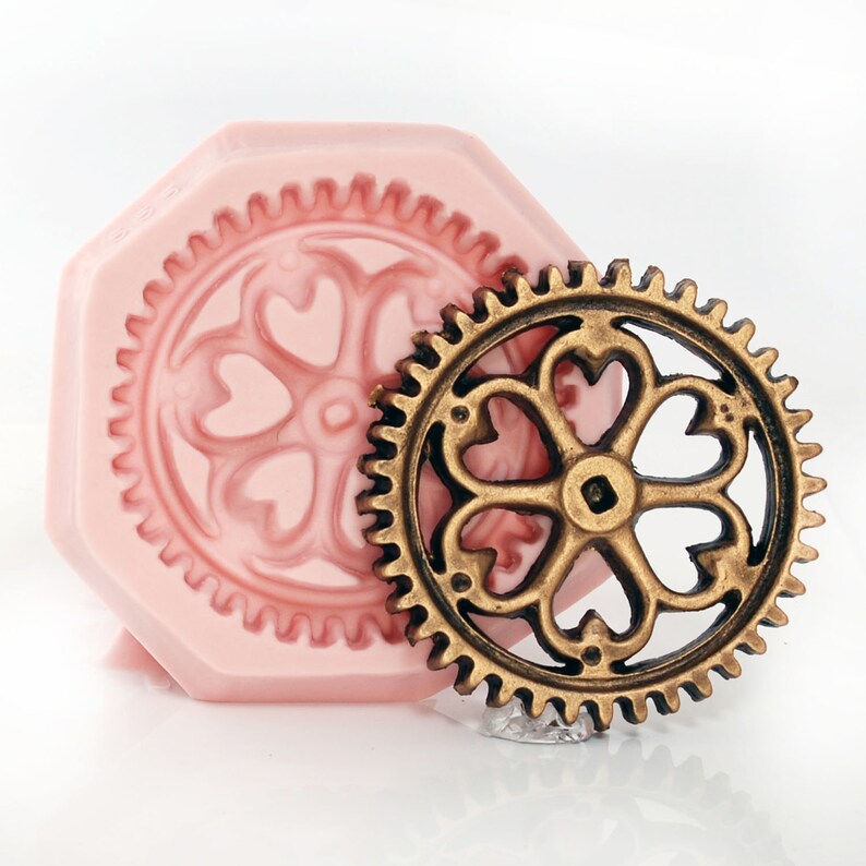 May include: A pink silicone mold with a detailed gear design featuring a five-pointed flower with heart-shaped petals. A gold metal gear with a similar design is placed next to the mold.