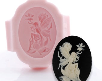 May include: A light pink silicone mold featuring an oval cavity with a fairy sitting on a flower design, paired with a finished black oval cameo piece displaying a raised white relief of the same fairy and flower motif.