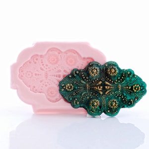 May include: A pink silicone mold with a detailed design of a green and gold decorative piece. The mold is used to create a replica of the decorative piece.
