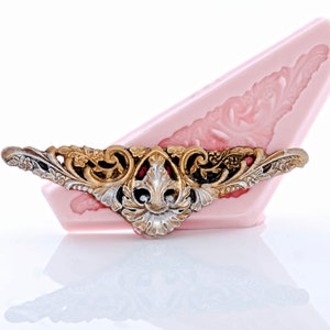 May include: A gold and silver decorative mold for making cake decorations. The mold is shaped like a stylized floral design with intricate details.