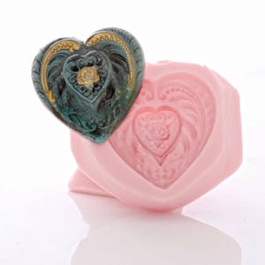 May include: A pink heart-shaped silicone mould with a detailed design. A green and gold heart-shaped charm is placed on top of the mould.