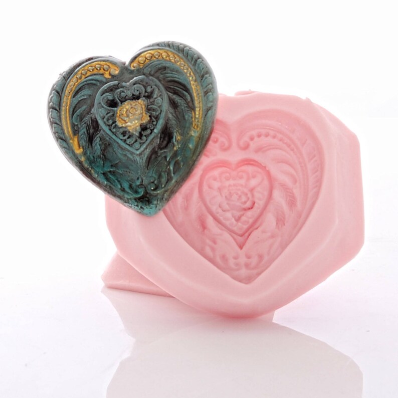 May include: A pink heart-shaped silicone mould with a detailed design. A green and gold heart-shaped charm is placed on top of the mould.