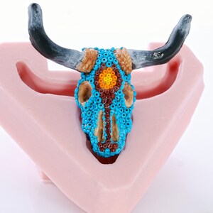 May include: A pink silicone mold shaped like a bull's head with a beaded bull skull inside. The skull is decorated with blue, brown, orange, and yellow beads.