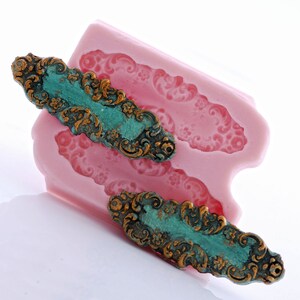 May include: Two ornate, antique-looking metal embellishments with a teal green patina and gold accents. The embellishments are sitting on a pink silicone mold.
