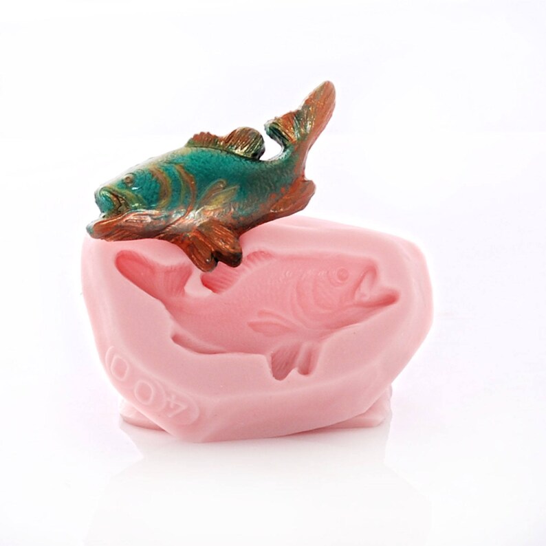 May include: A pink silicone mold with a raised fish design. A small, green and copper colored fish is sitting on top of the mold.