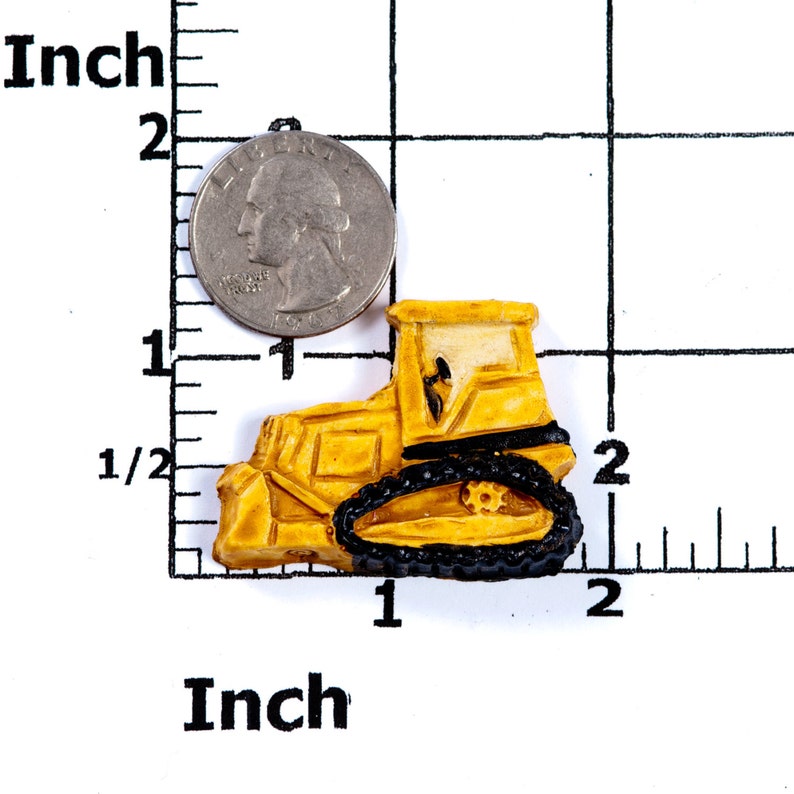 Puede incluir: Un bulldozer de juguete amarillo con orugas negras, que mide aproximadamente 2,5 cm de largo. El bulldozer est&aacute; sobre una superficie blanca con una regla para la escala.