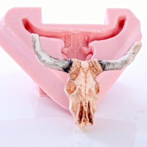 May include: A pink silicone mold for making a resin bull skull. The mold is open and the bull skull is visible inside. The bull skull is detailed with horns, eyes, and a textured surface.