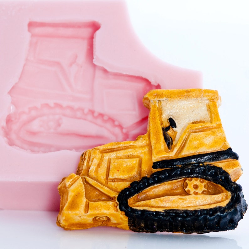 Puede incluir: Un bulldozer de juguete amarillo con orugas negras, sentado en un molde de silicona rosa. El bulldozer tiene un dise&ntilde;o detallado con una cabina del conductor y una cuchilla levantada.