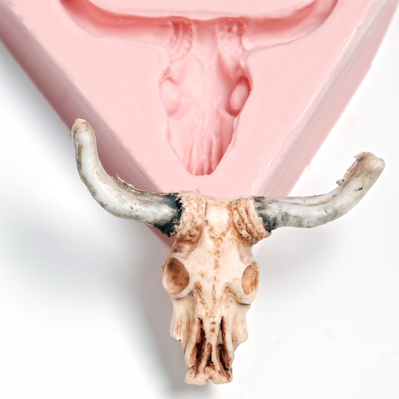 May include: A white ceramic bull skull with horns, sitting on a pink silicone mold. The skull has detailed features and a textured surface.
