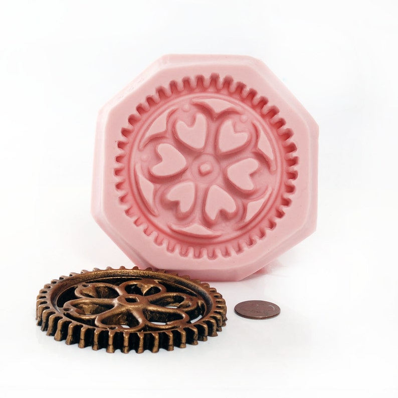 May include: A pink silicone mold with a detailed design of a flower with heart-shaped petals. A bronze metal gear with a similar flower design is also shown.