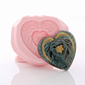 May include: A pink silicone heart-shaped mould with a teal and gold heart-shaped ornament inside. The ornament has a detailed design with a floral pattern.