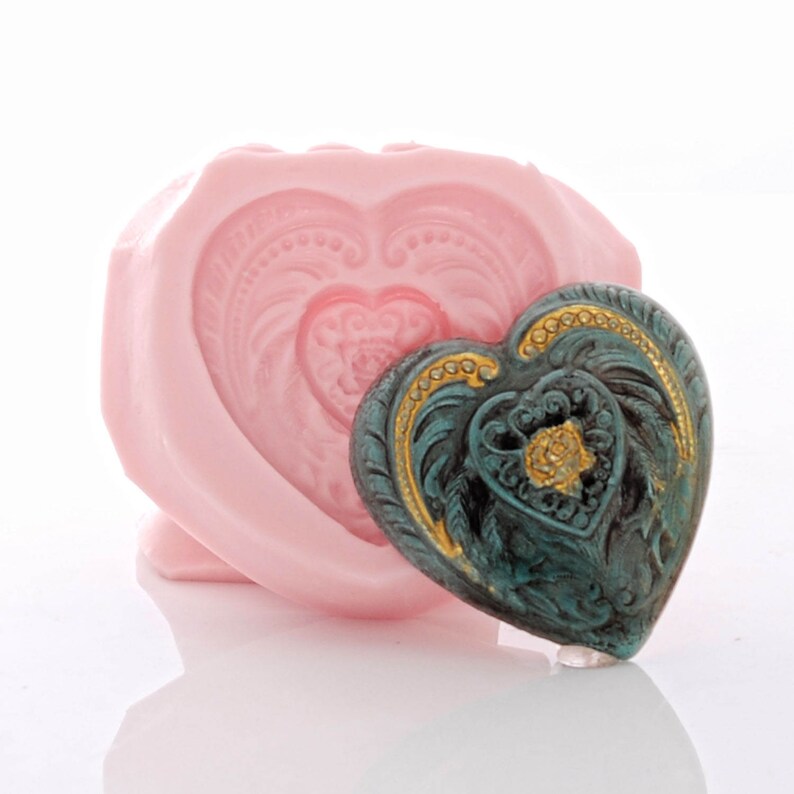 May include: A pink silicone heart-shaped mould with a teal and gold heart-shaped ornament inside. The ornament has a detailed design with a floral pattern.