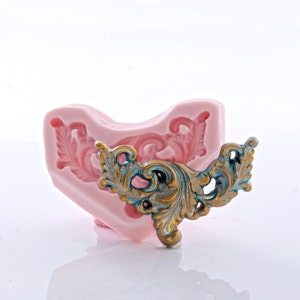 May include: A pink silicone mold with a detailed floral design, next to a finished decorative piece. The decorative piece is gold and teal, with intricate scrollwork and a vintage aesthetic. The mold is heart-shaped.
