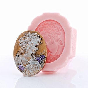 May include: An oval-shaped silicone mold in light pink featuring a recessed cameo design of a person with flowers. Beside it sits a finished cameo plaque in metallic gold and silver tones, depicting a person with purple floral accents.